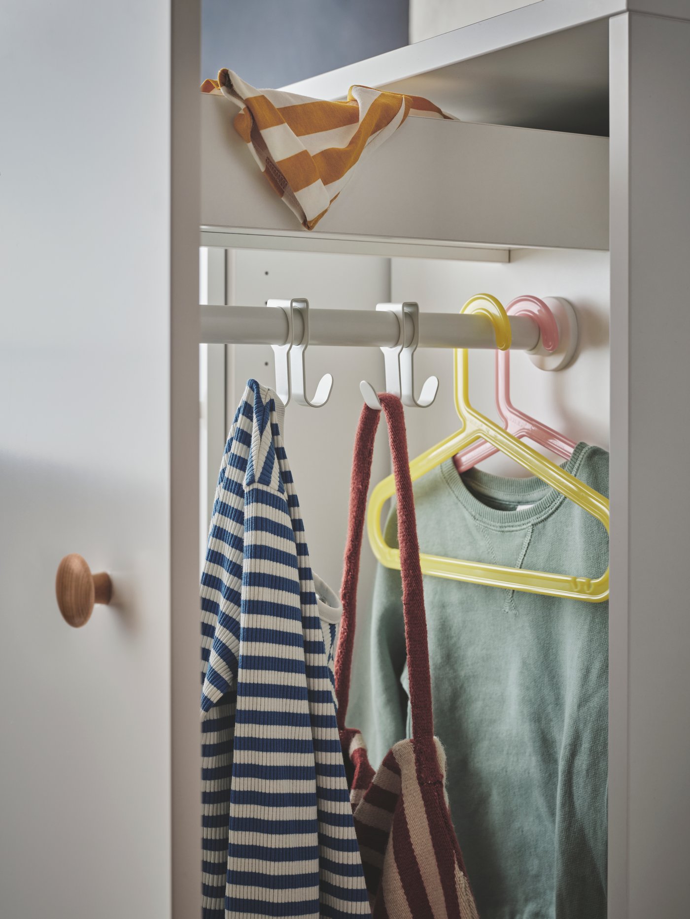 The white SMÅSTAD pull-out storage unit with a top drawer and hooks and hangers holding children’s clothes and a bag.