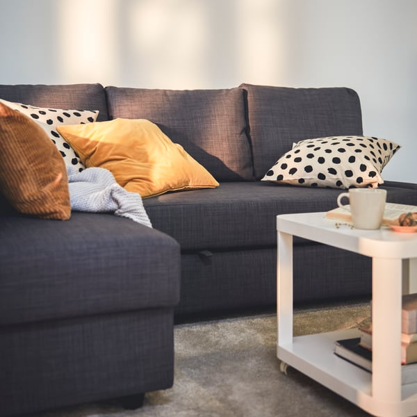 Scattered sunlight over a dark-grey FRIHETEN sofa-bed with a throw and different-colour cushions strewn across it.