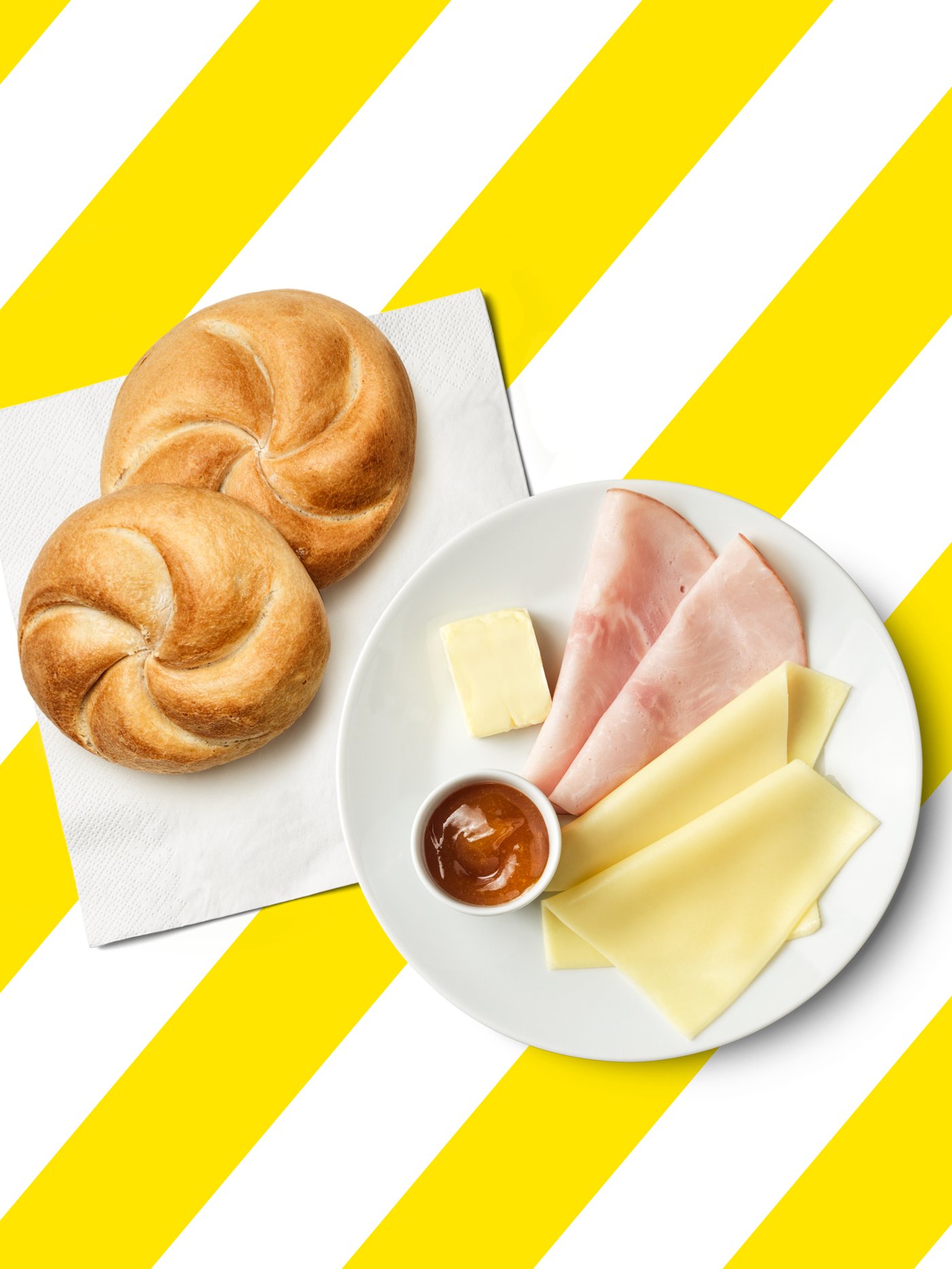 Ein Teller mit Schinken, Käse, Marmelade und Butter, daneben zwei Semmeln vor einem gelb-weiß gestreiftem Hintergrund. 