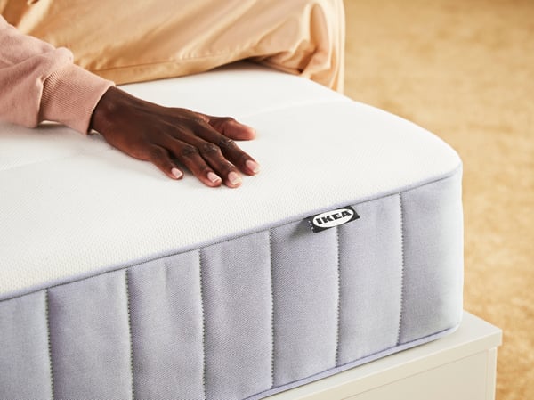 A person sitting near the corner of a firm/light blue VESTERÖY pocket sprung mattress with their hand on the surface.