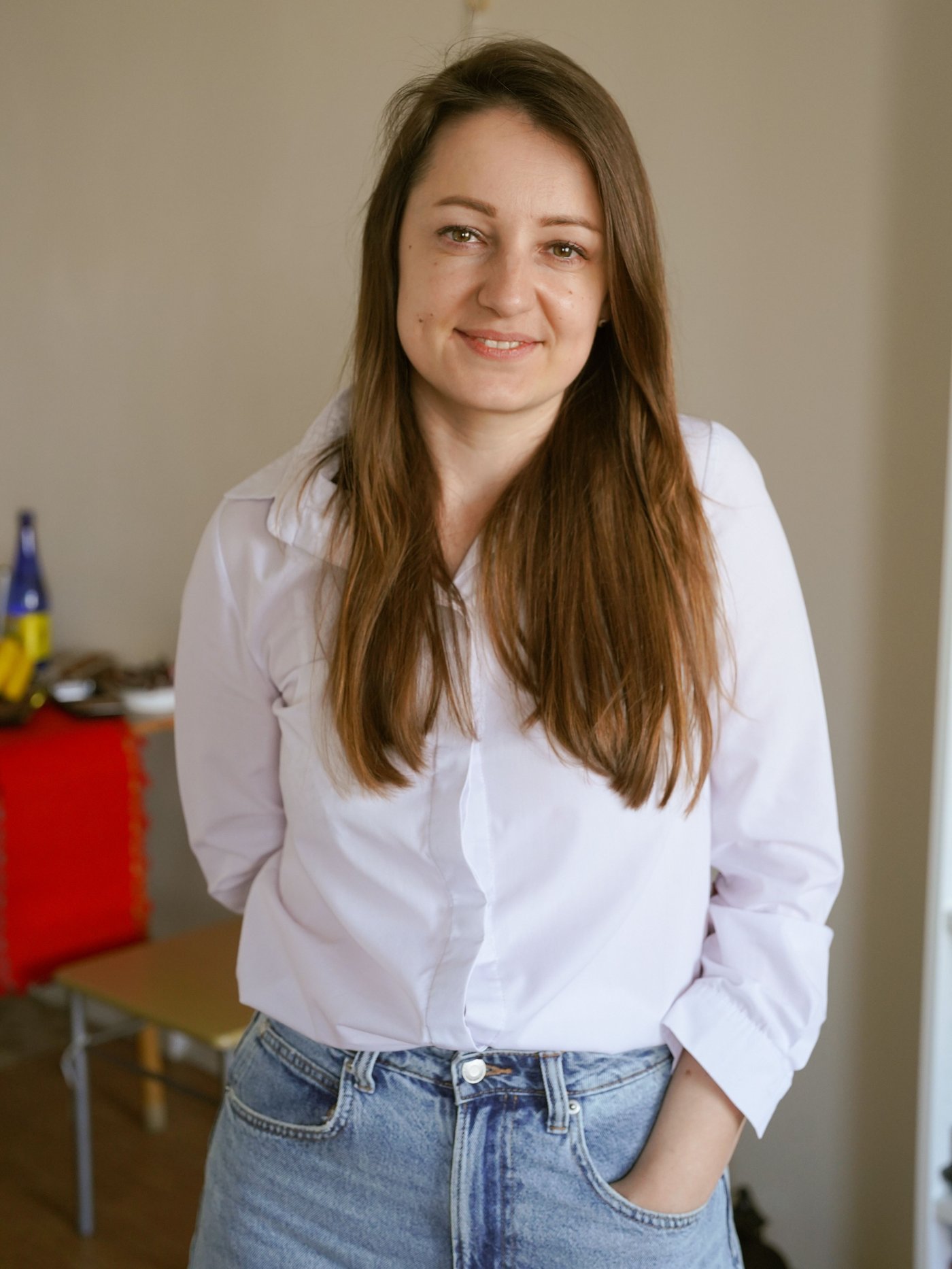 Dívka v bílé košili s hnědými rozpuštěnými vlasy stojí uvnitř místnosti a dívá se do objektivu fotoaparátu.