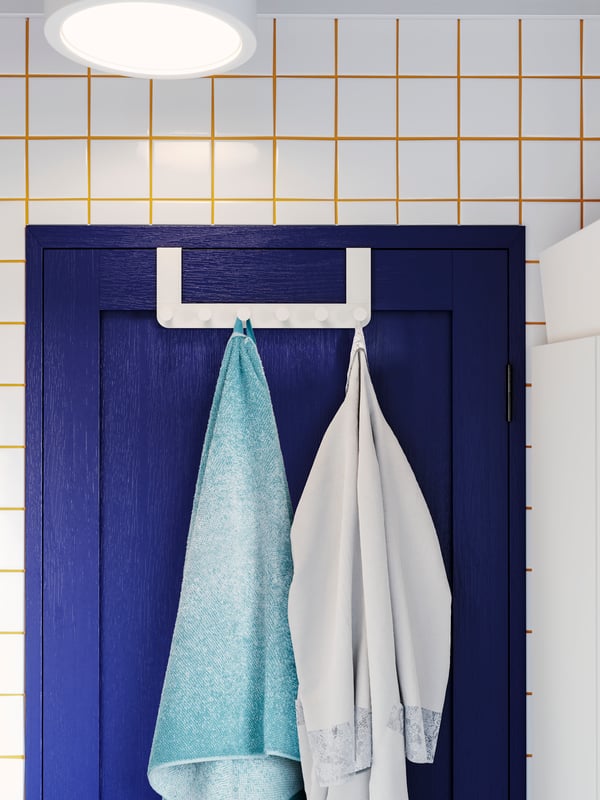 A white hanger for door hung over a blue door, with a towel and a bathrobe on the hooks. White tiles and yellow grout.