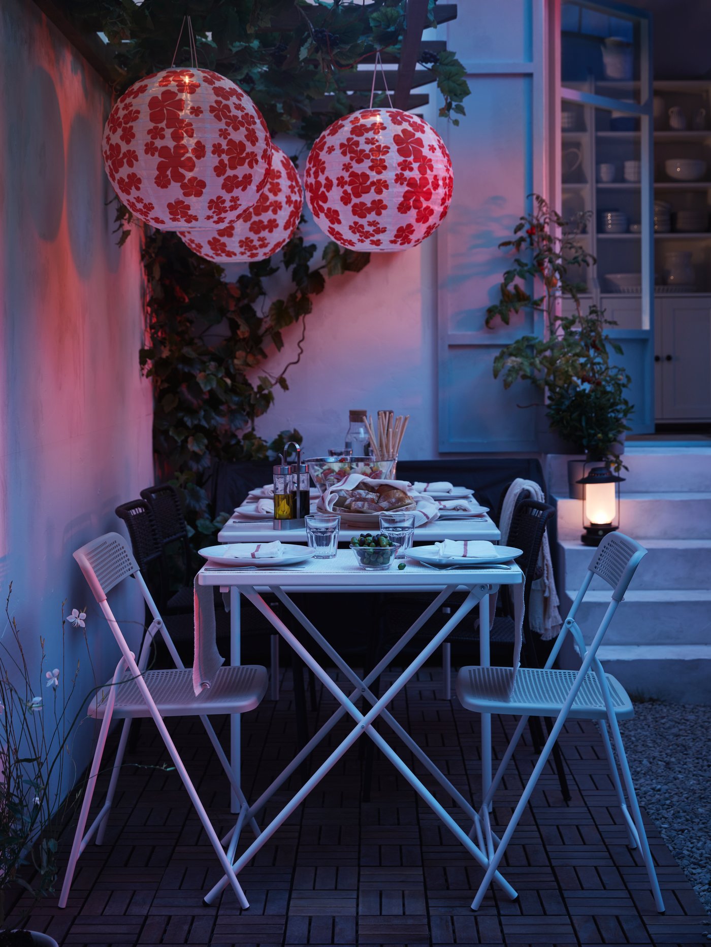 Una mesa de comedor exterior TÖRPARÖ grande junto a una pequeña y plegable ubicadas en un jardín con piso RUNNEN. Frente a la mesa pequeña, hay dos sillas plegables TÖRPARÖ blancas. El conjunto está iluminado por tres lámparas de papel SOLVINDEN en colores blanco y rojo. 