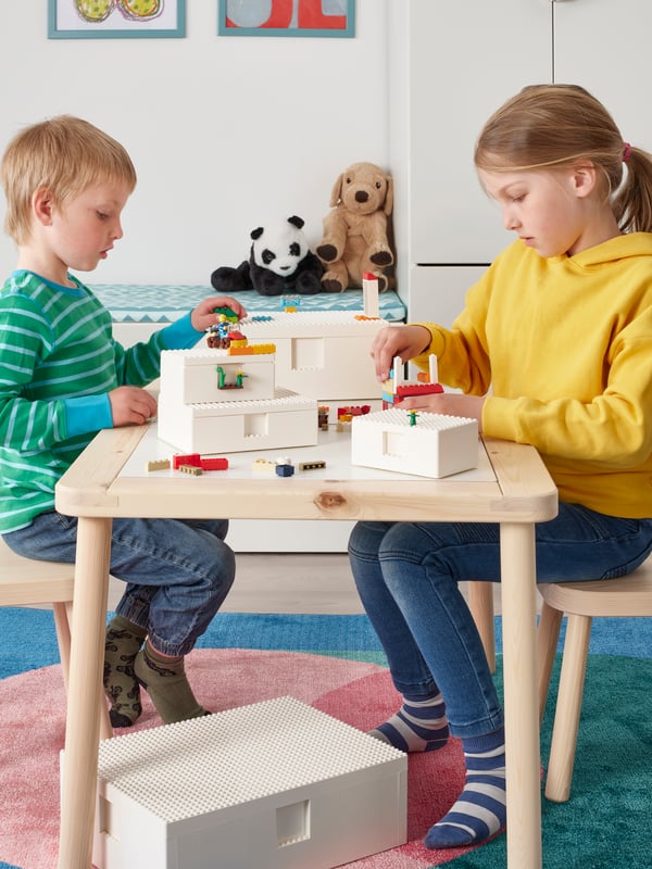 Two children play with LEGO bricks together at a wooden table, attaching LEGO minifigures to two white BYGGLEK boxes.