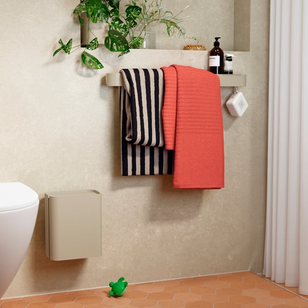 A bathroom with a beige FLORSJÖN waste bin placed next to folded towels and a green plant on the wall shelf.
