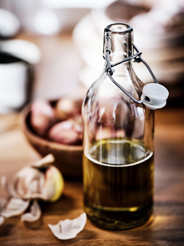 Une bouteille en verre avec un bouchon sur une surface en bois, à moitié remplie d’huile d’olive, à côté d’un bol rempli de têtes d’ail.