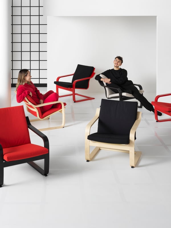 Six POÄNG low-back armchairs in different colours and cushions are in a white room. Two people are socialising in them.