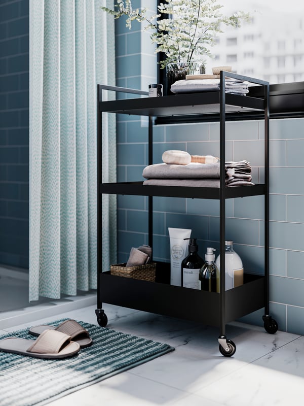 A black NISSAFORS trolley with shampoo bottles and towels stored on it is standing next to a shower in a turquoise bathroom.