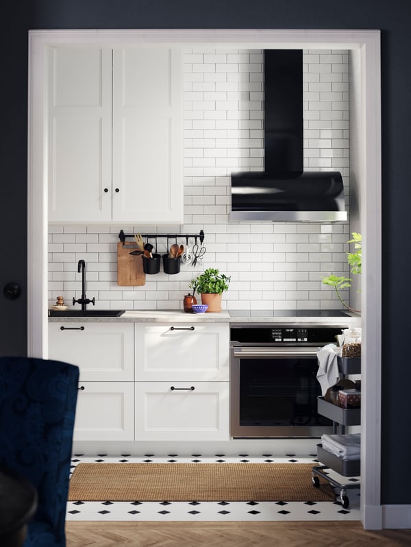 A kitchen with AXSTAD matte white kitchen cabinet doors with black handles, a black tap, a trolley, against a tiled wall.