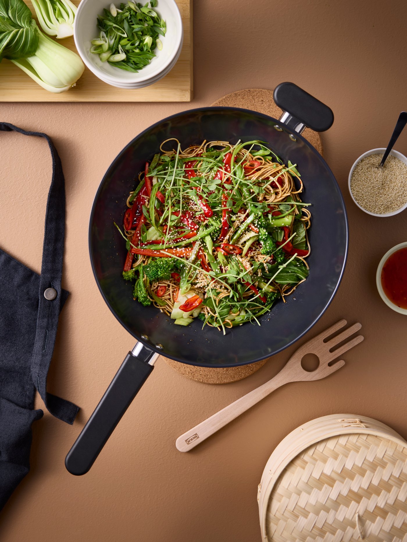 A MIDDAGSMAT wok in stainless steel with a non-stick coating, placed on HEAT pot stands, has vegetables and noodles inside.