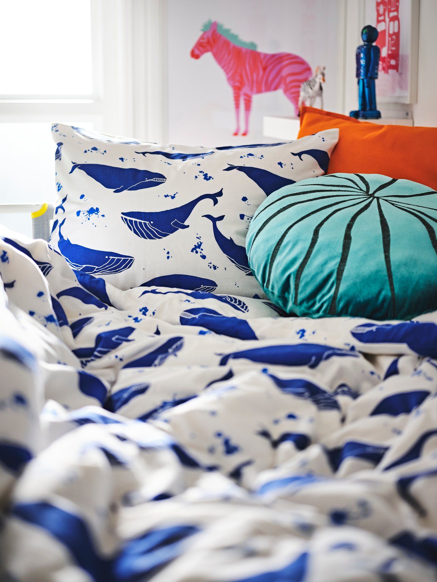 A child’s bed set up with BLÅVINGAD bedding and a cushion. A framed art print of a red and white zebra hangs beyond it.
