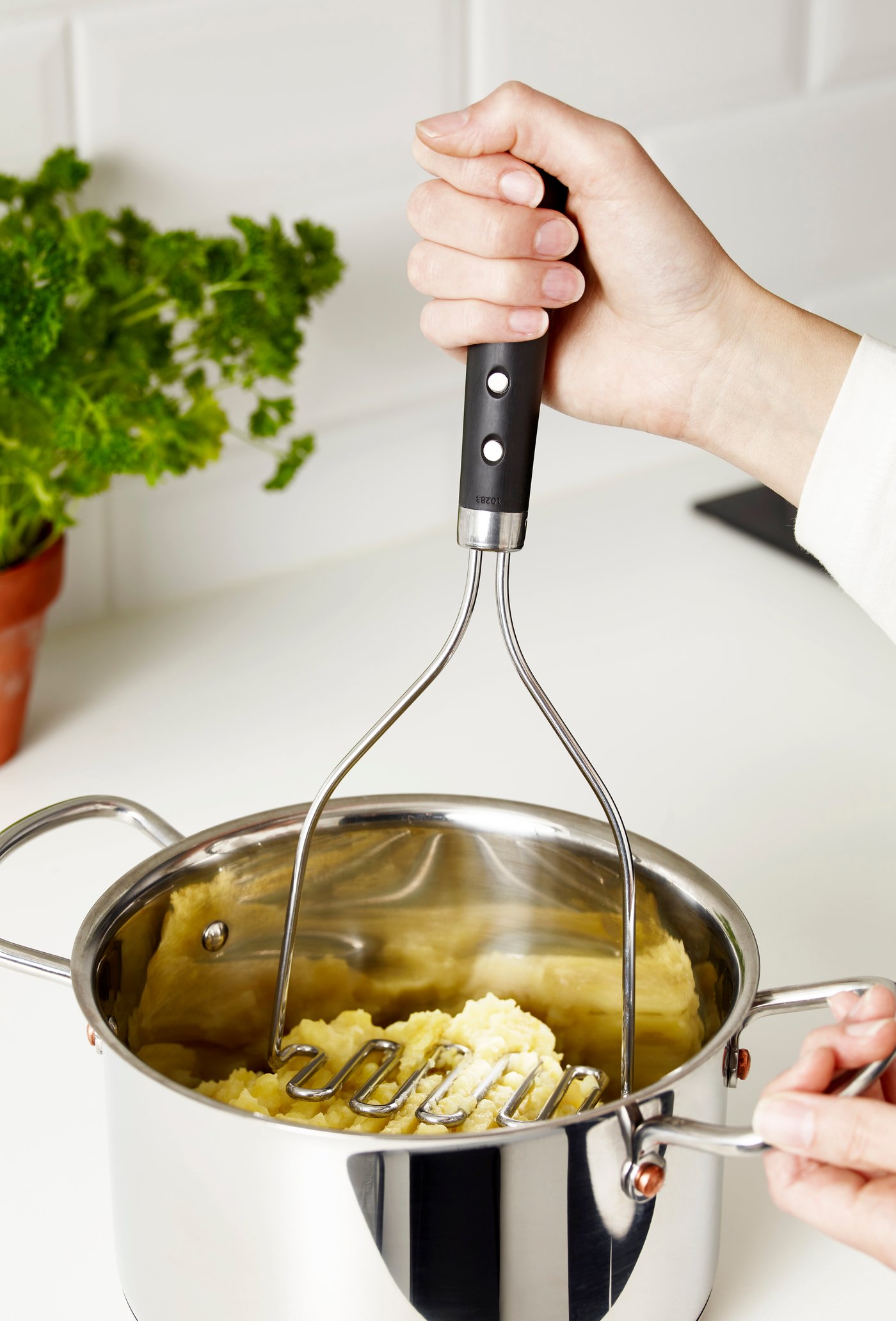 A hand is holding a VARDAGEN potato masher, mashing boiled potatoes in a pot made of stainless steel on a white surface.