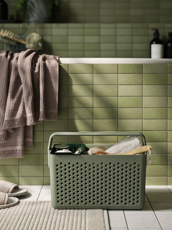Una canasta de almacenamiento UPPRÄMEN en verde grisáceo lleno de artículos de cuidado corporal en el suelo de un baño delante de una bañera verde.
