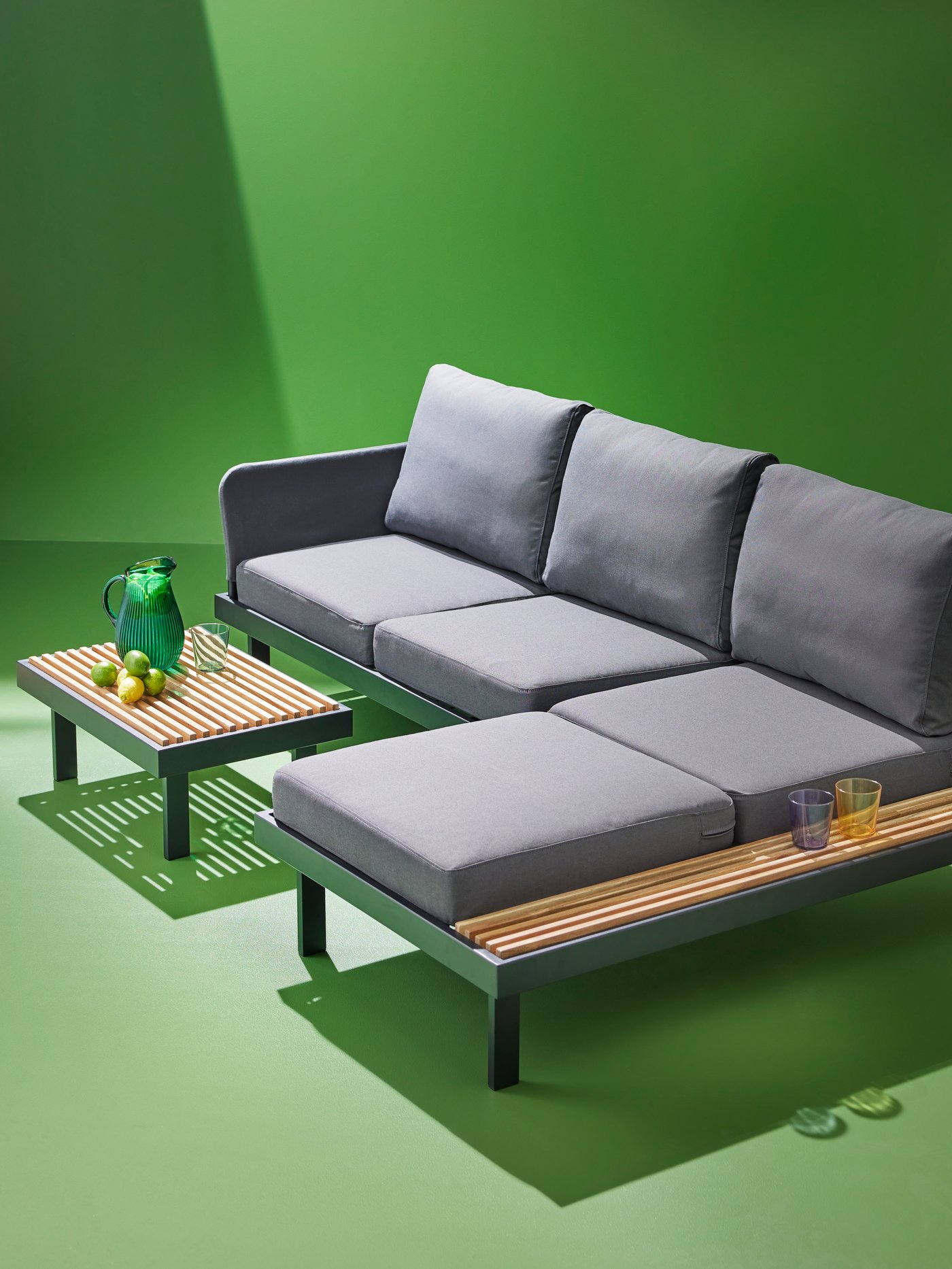 A REVSKÄR 3-seat conversation set, in anthracite, in a green space. There is a green jug, glass and limes on the side table.