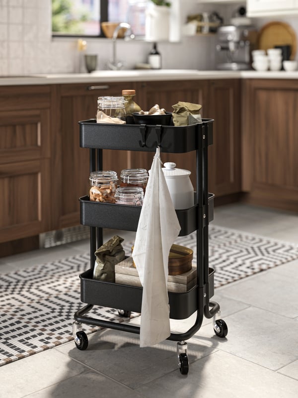 Un carrito RÅSKOG de color negro con botellas de vidrio SÄLLSKAPLIG, tazas GLADELIG, vasos POKAL, frascos y paños de cocina. 