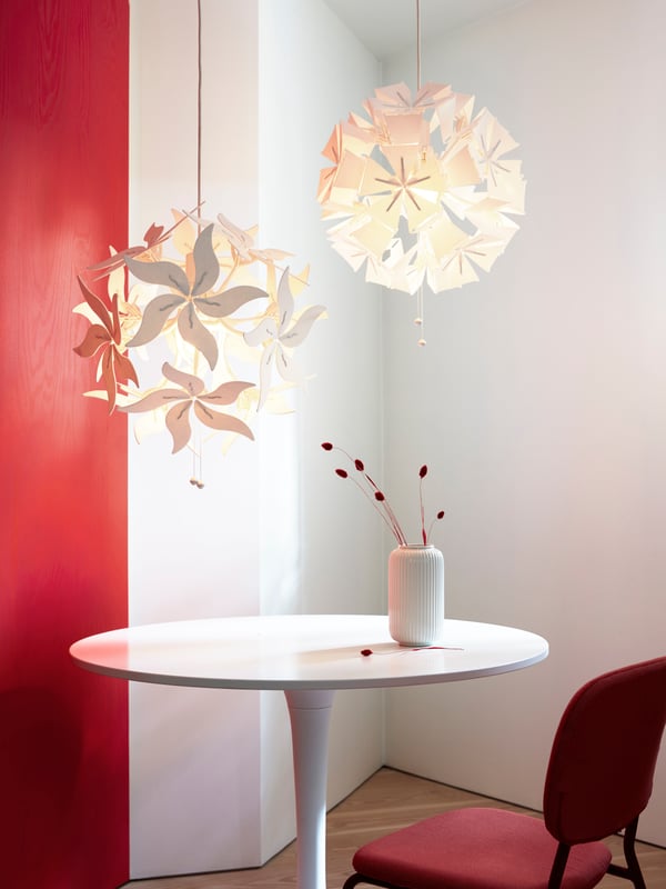 Una mesa redonda blanca con una silla KARLJAN roja, arriba de la mesa hay dos lámparas de techo RAMSELE en forma de flor encendidas.