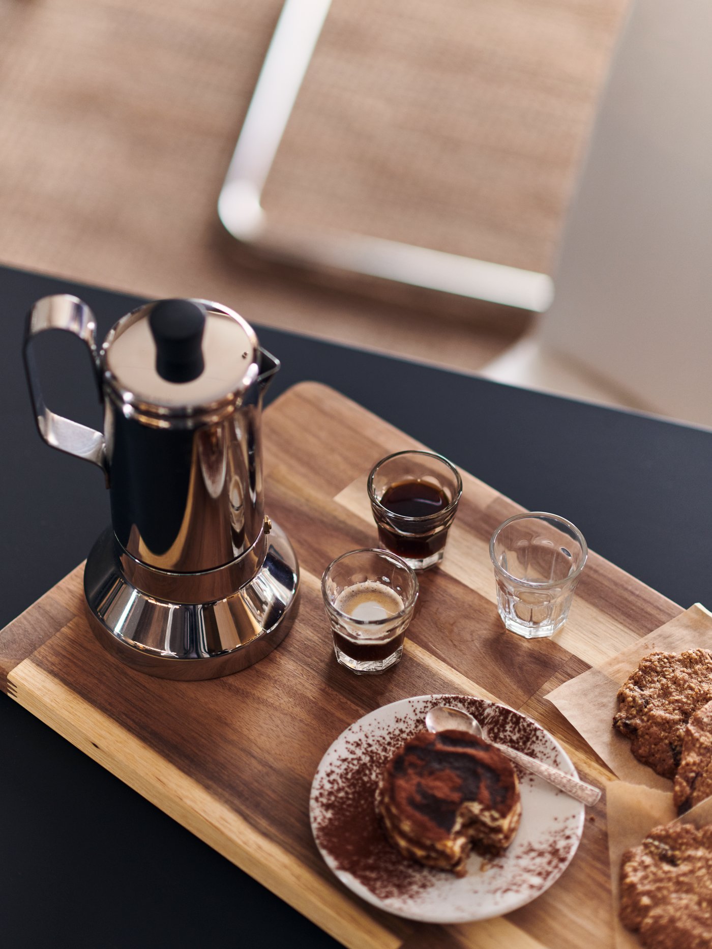 A METALLISK espresso maker on a SMÅÄTA chopping board beside biscuits, a cake, and small POKAL glasses filled with espresso.