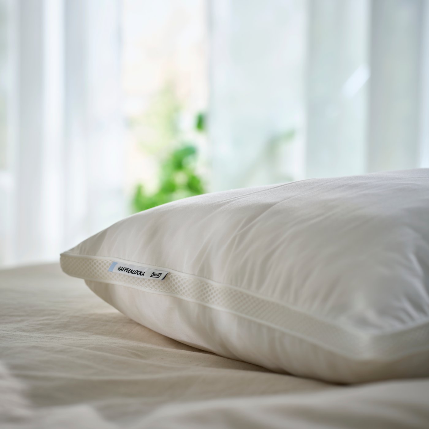 GAFFELKLOCKA pillow in white placed on a neatly made bed with soft beige bedding beside a bright window.