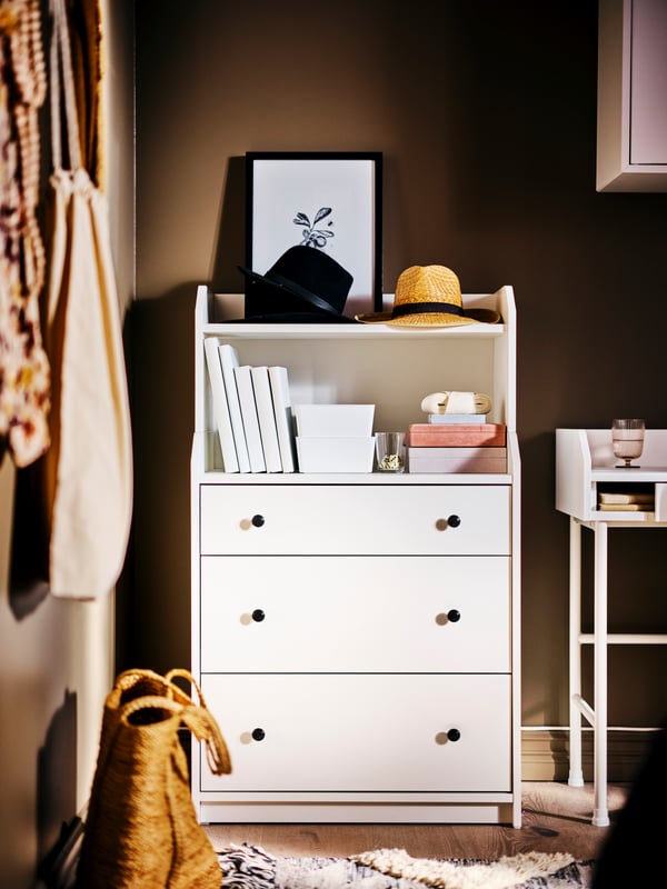 Un mueble de almacenaje HAUGA blanco, sobre el cual hay varios libros, cajas, sombreros y un cuadro de adorno.