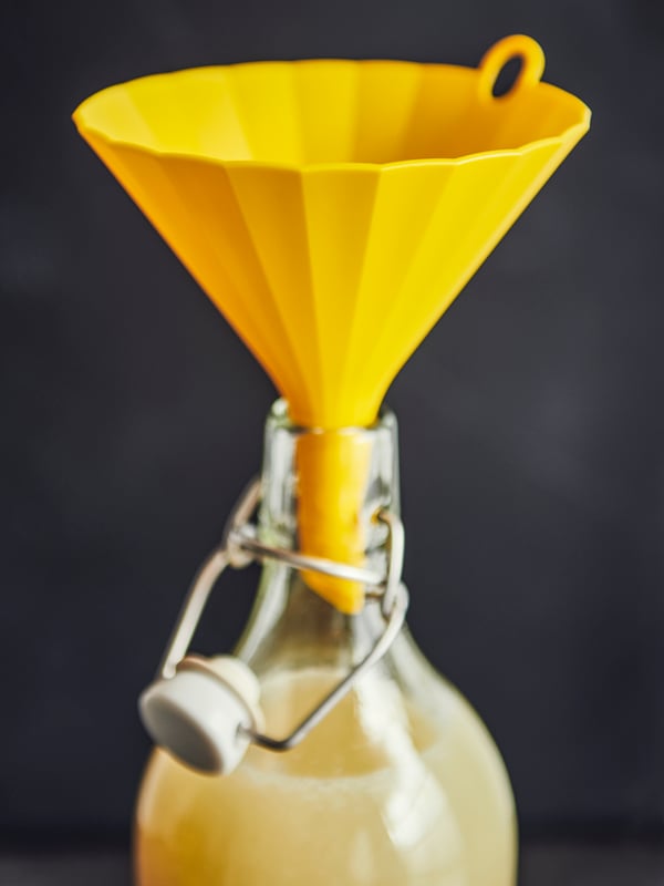An UPPFYLLD funnel in bright yellow placed in the neck of a KORKEN bottle with stopper in clear glass, liquid in the bottle.
