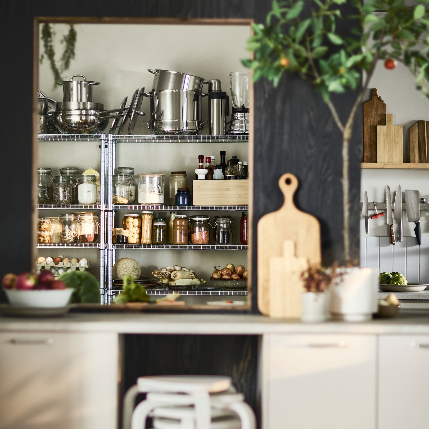 A warm and well-organised IKEA kitchen features metal shelving filled with jars, spices, pantry goods and stainless-steel utensils, while chopping boards, knives and plants add a natural touch.