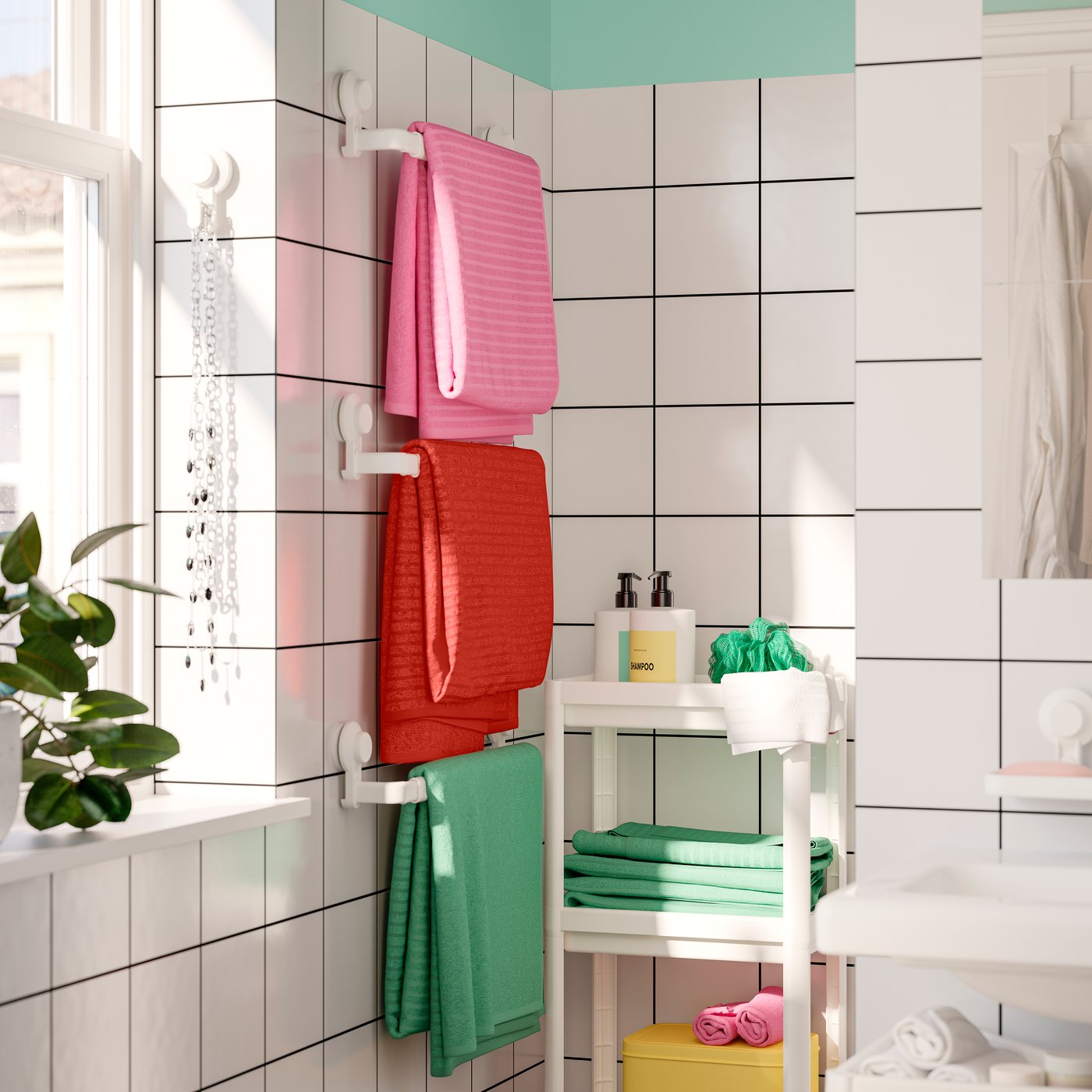 Three VÅGSJÖN towels in pink, red and green hang on white TISKEN towel racks in a white bathroom. A plant is by the window.
