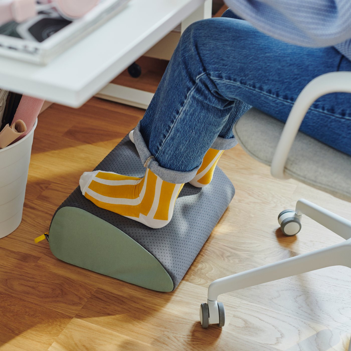 Eine Person sitzt an einem weißen Schreibtisch und ihre Füße ruhen auf einer ergonomischen ÖVNING Mehrzweck-Fußstütze.