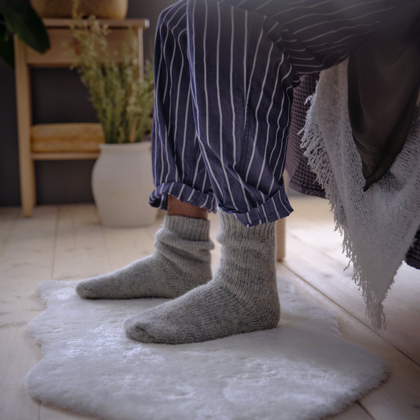A close-up of a person seated on the edge of a bed. They are wearing grey socks and their feet are on a white TOFTLUND rug.
