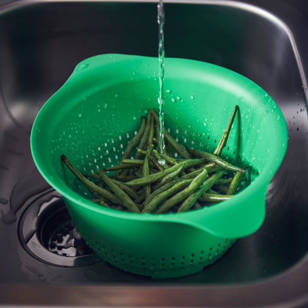 An UPPFYLLD colander in bright green sitting in a sink, the colander contains green beans and there is water pouring in.