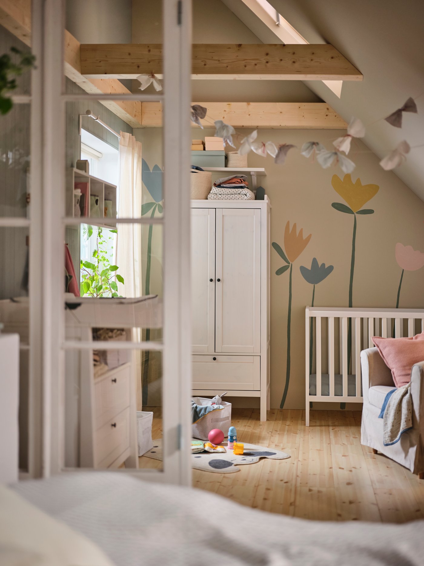 A white SUNDVIK wardrobe in a baby’s bedroom beside a white SUNDVIK cot. A flower mural is on the wall behind them.