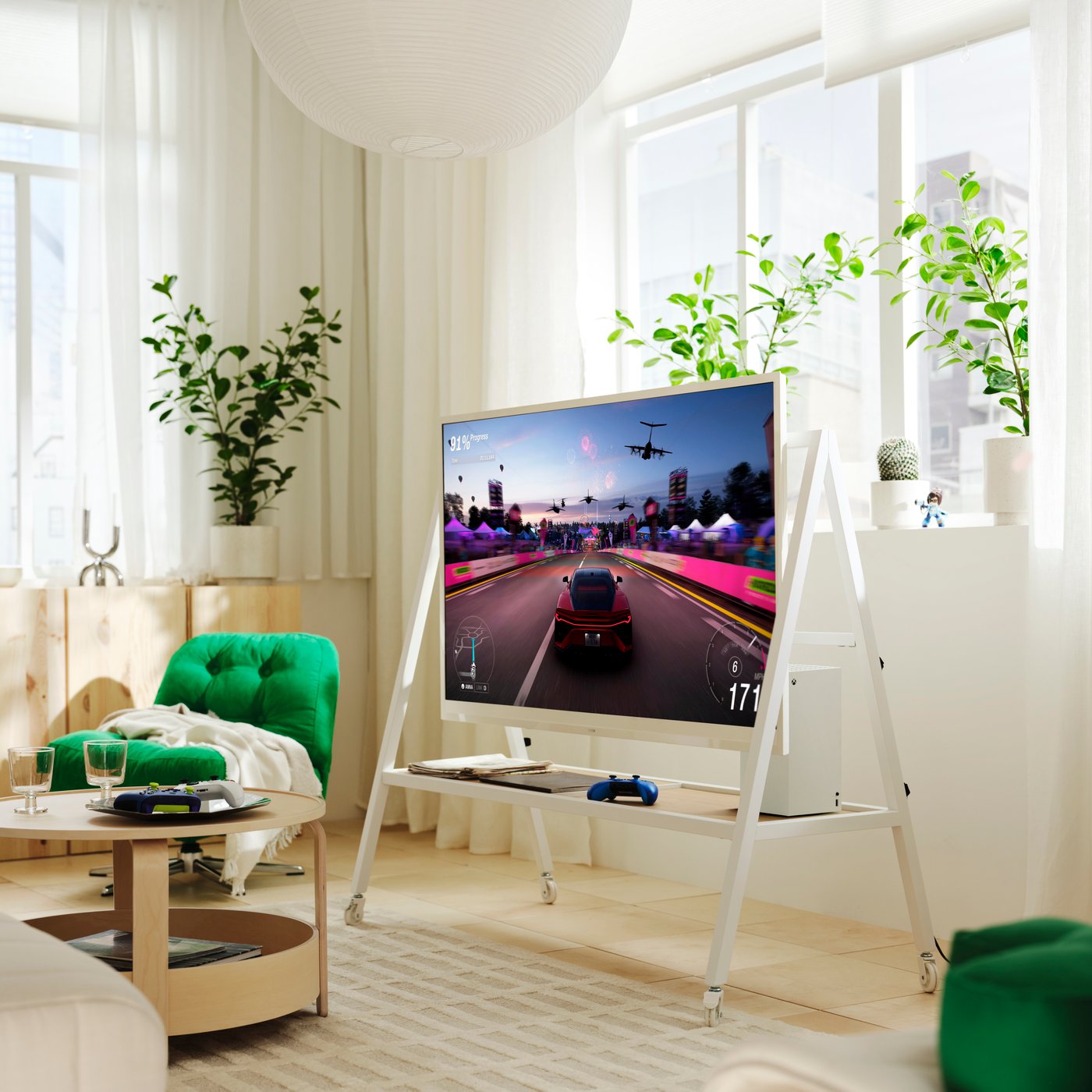 A white YXSTABY gaming TV stand on castors is placed in front of a window in a light living room. The TV shows a car game.