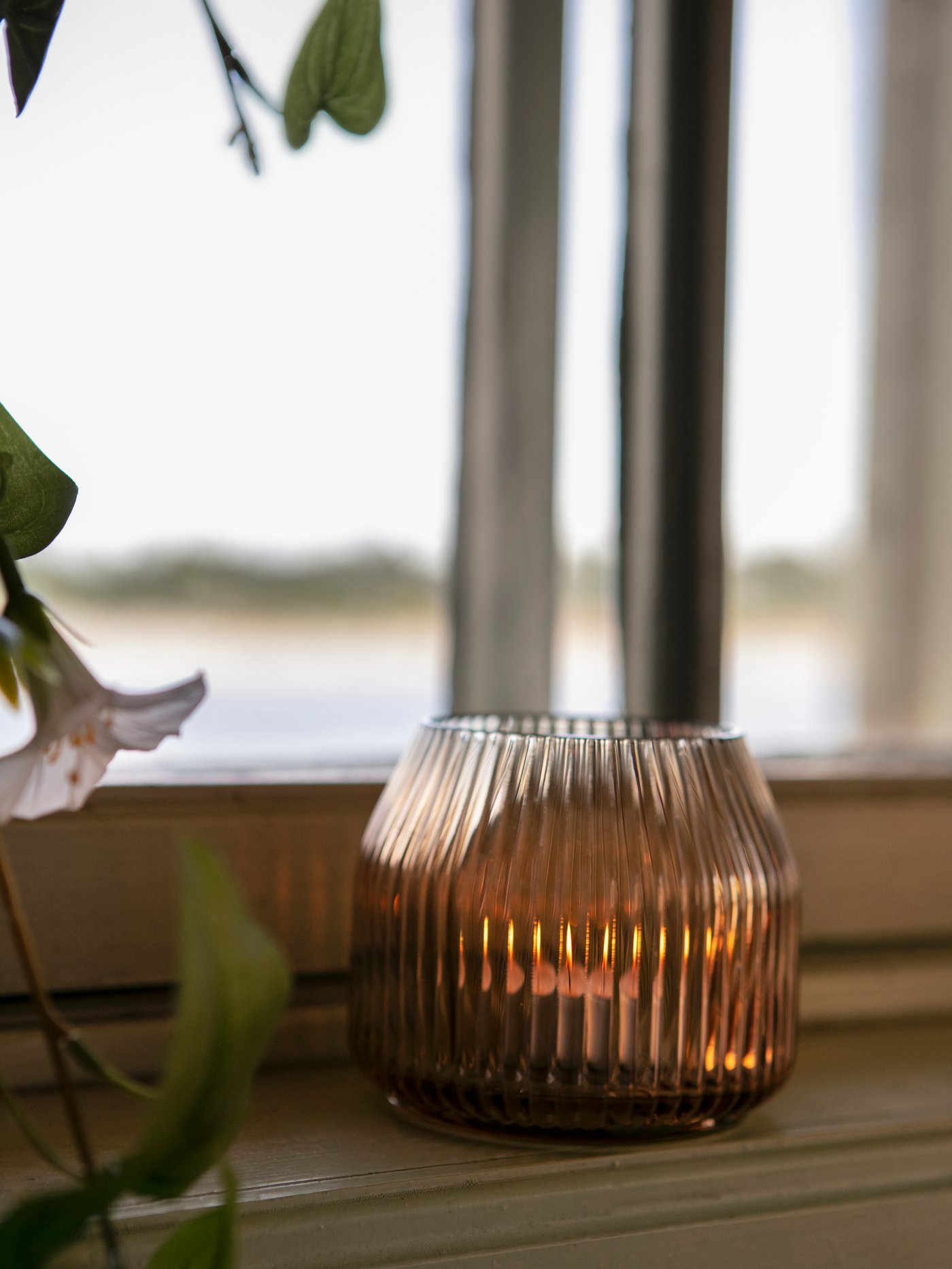 An ANLEDNING tealight holder with a ridged pattern in light brown glass with a lit candle inside standing by a window.