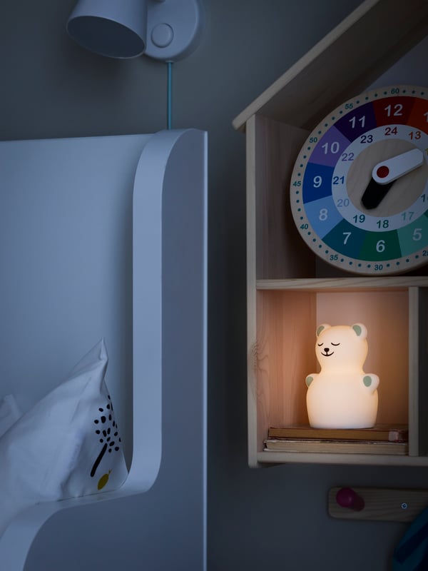 Tamna soba s TÖVÄDER LED noćnim svetlom u obliku medveda u kući za lutke/zidnoj polici pored belog produživog kreveta i zidne lampe.
