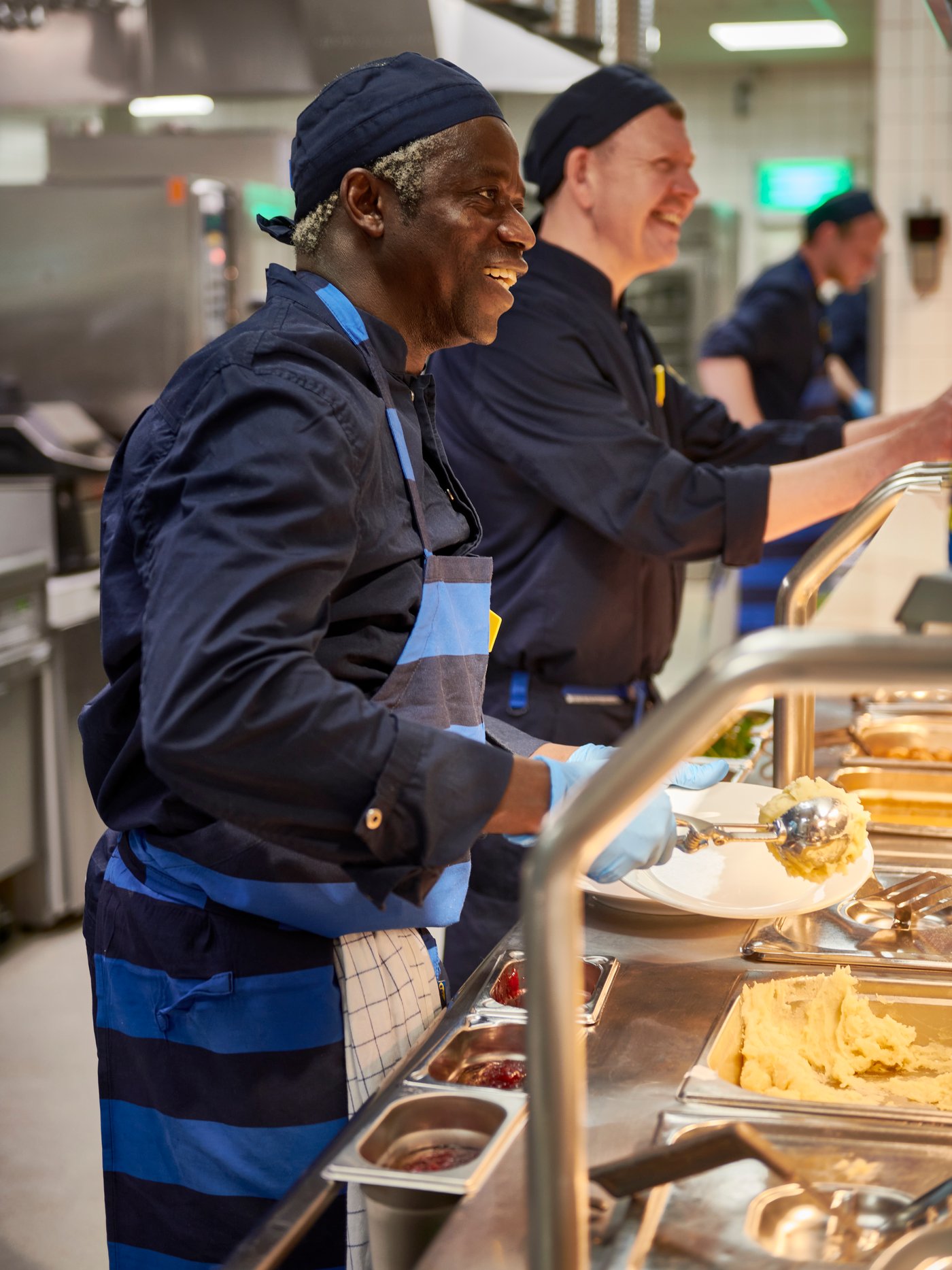 IKEA-Küchenmitarbeiter in dunkelblauer Uniform mit blauer Schürze serviert lächelnd frisches Essen in der IKEA-Restaurantküche. Neben ihm steht ein Kollege und im Vordergrund sind Edelstahltabletts mit Speisen zu sehen