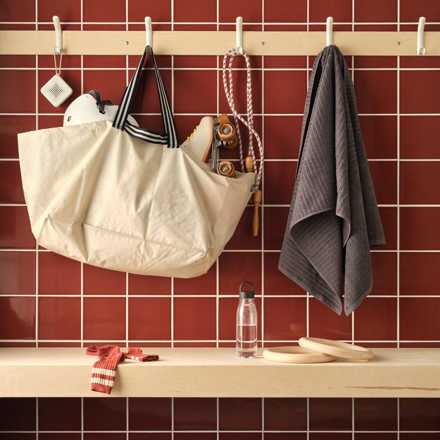 Eine geflieste Wand mit einer GÖRSNYGG Tragetasche und einem VÅGSJÖN Handtuch an Haken sowie einer IKEA 365+ Trinkflasche auf einer Bank.