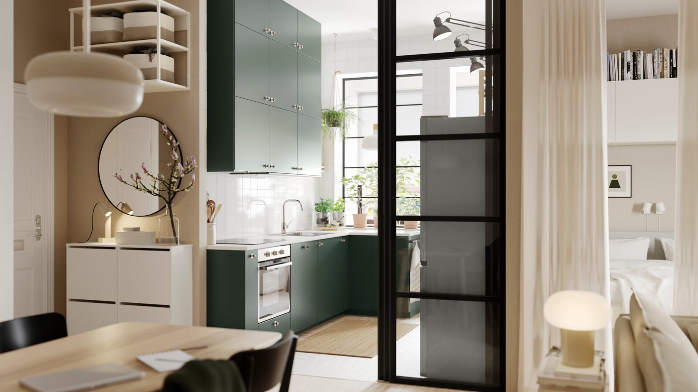 An open kitchen with deep green HAVSTORP kitchen fronts and wall cabinets up to the ceiling, next to a bright living room.