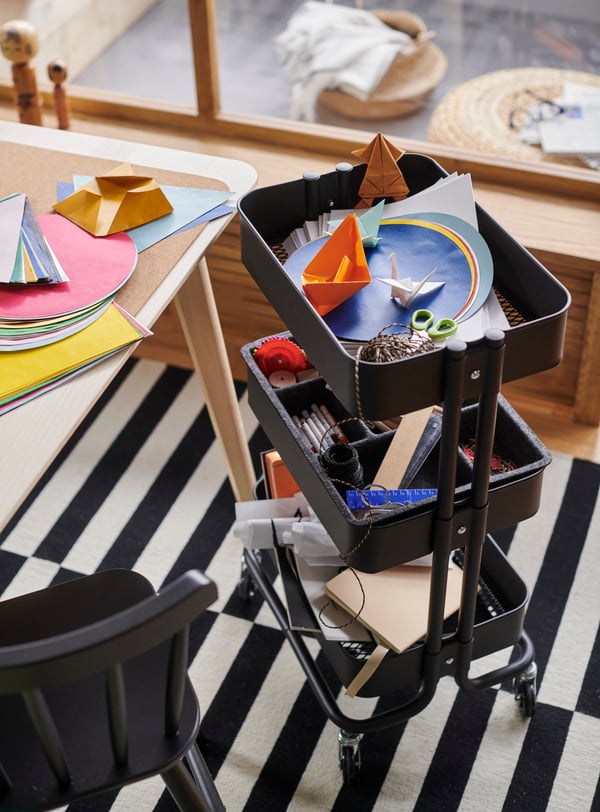 Black RÅSKOG trolley laden with arts-and-crafts accessories placed next to a table top covered with scattered coloured paper.