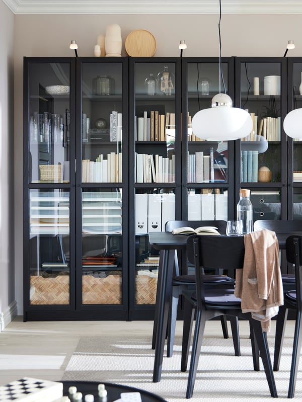 A bright room with three tall glass-door bookcases in front of a LISABO dining table with two DEJSA pendant lamps above it.