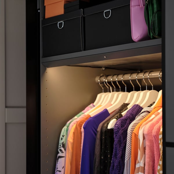 Kleidung an BUMERANG Kleiderbügeln hängt in einem PAX/MEHAMN Kleiderschrank unter einem Boden, auf dem sich schwarze NIMM Kästen mit Deckel befinden.