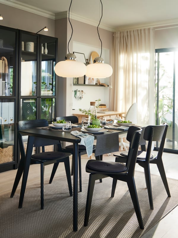 A black LISABO dining table with four chairs around it laid with plates, bowls and glasses for a meal, in a bright room.