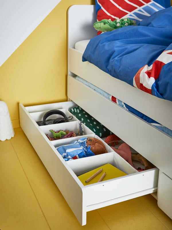 A white children's bed with storage underneath revealing colouring materials, toys, bed linen, headphones in tidy boxes.