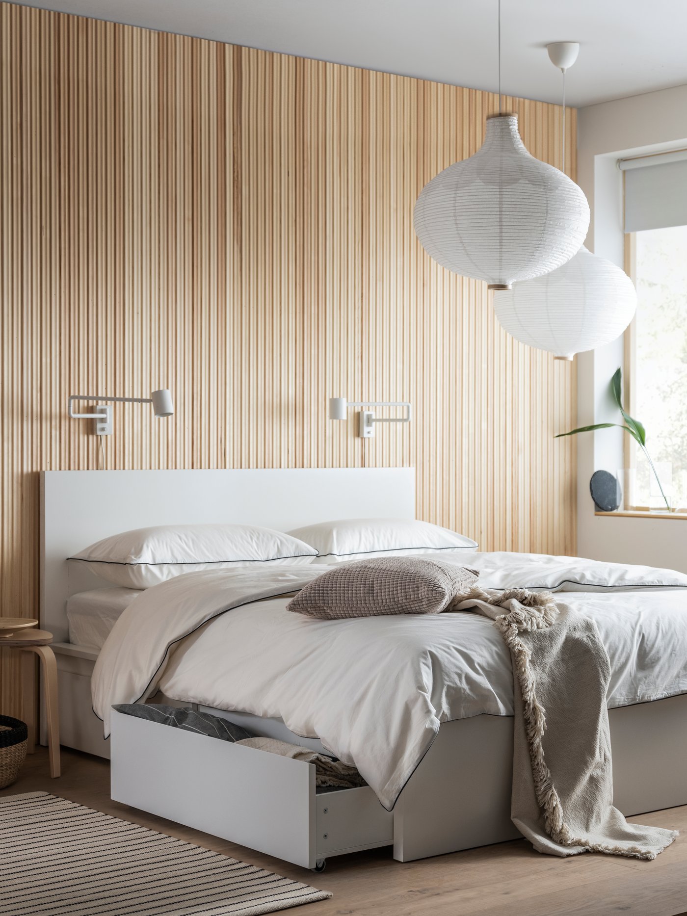 A white MALM bed with storage covered with KUNGSBLOMMA bed linen, with one storage box open and two RISBYN lampshades above.