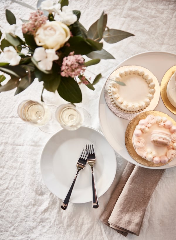 Un dessus de table avec des gâteaux de mariage miniatures sur un plat à gâteau UPPLAGA, des verres à champagne STORSINT, de la vaisselle et des décorations.