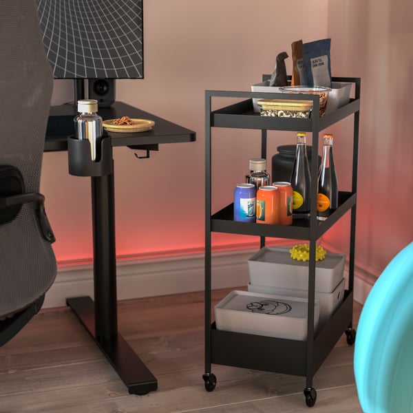 Neben einem Gamingschreibtisch steht ein NISSAFORS Servierwagen in Schwarz. Auf den Böden des Servierwagens sind Snacks, Getränke und Boxen angerichtet.