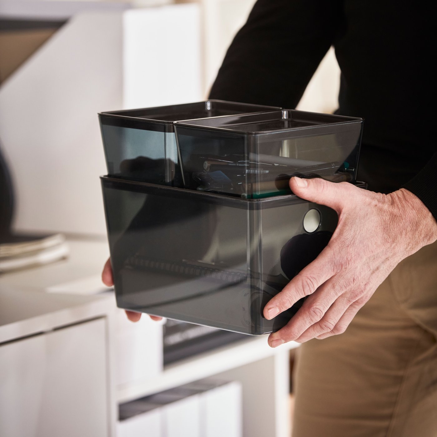 Close-up of a man holding three black KUGGIS storage boxes stacked together. A smaller box shows pens stored inside.