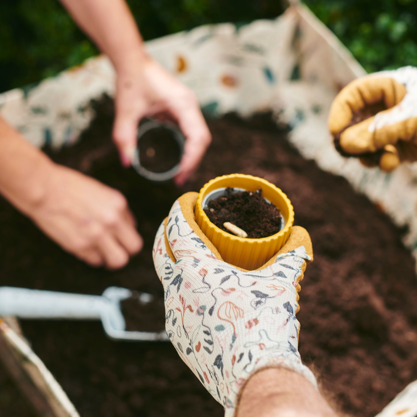 The hands of two people planting seeds in DAKSJUS plant pots next to a sprout-patterned DAKSJUS potting mat filled with soil.