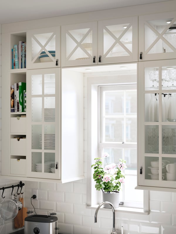 Off-white kitchen cabinets arranged around a window with off-white BODBYN vitrine glass doors with crossbars.
