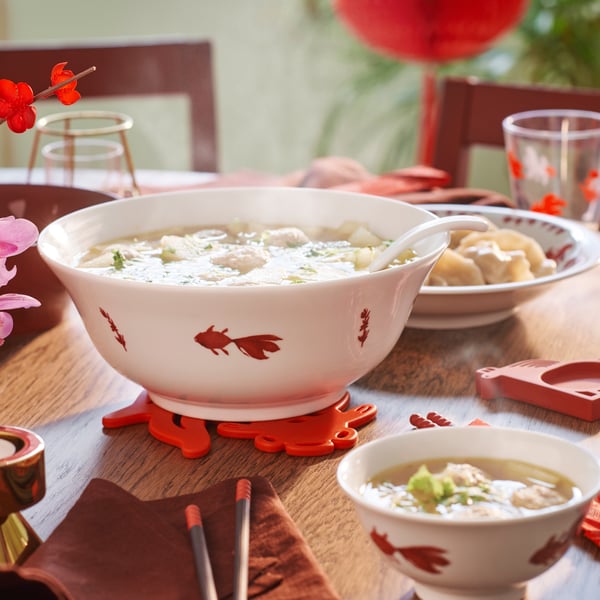 A white FÖSSTA bowl, decorated with red goldfish and filled with soup and dumplings, sits on a table with festive tableware.