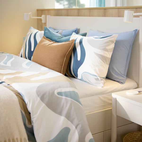 Ein Hotelbett, das neben einer niedrigen Trennwand steht, ist mit Bettzeug in MYGGLASVINGE Bettwäsche mit grossem Blattmuster bezogen.