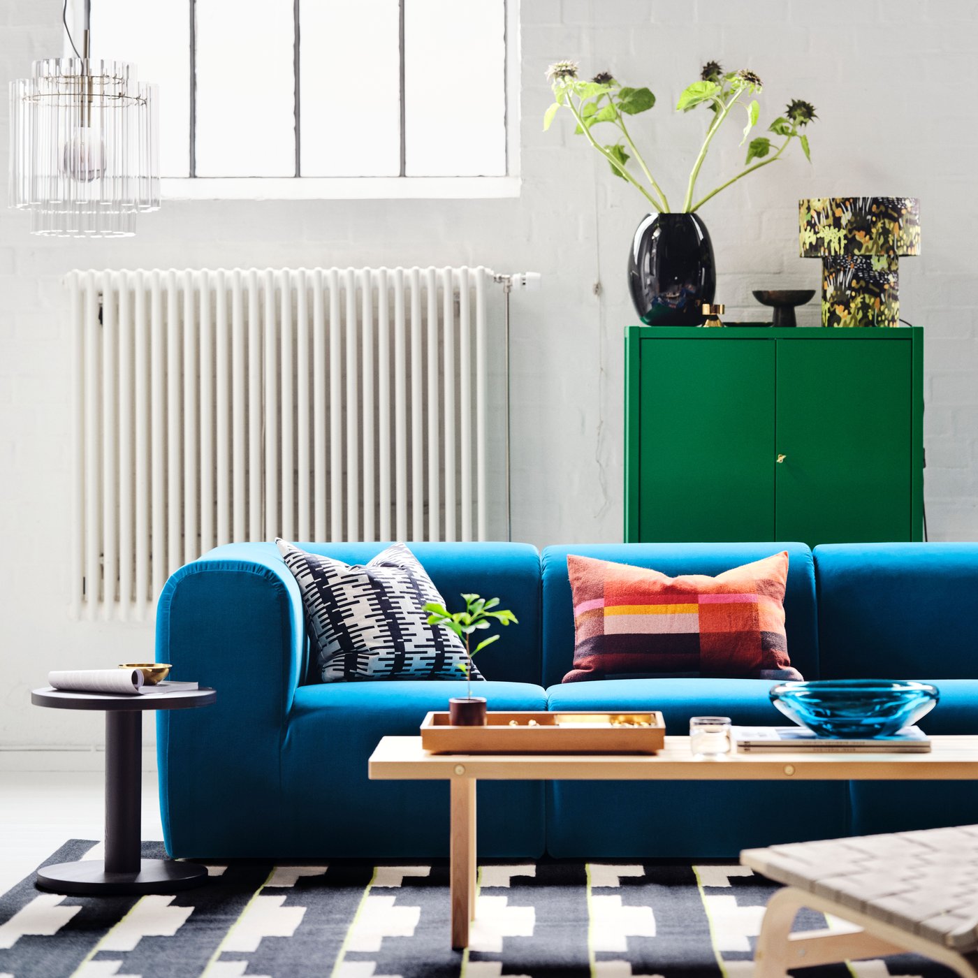A white room with a dark turquoise STOCKHOLM 2025 3-seat sofa, with two tables on a rug in front, and a green cabinet behind.